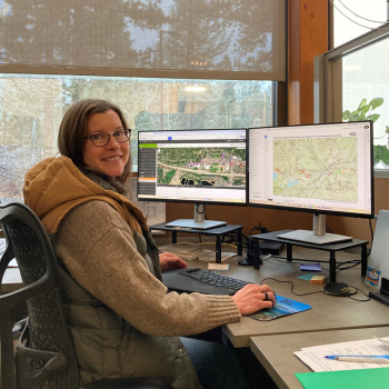 A women sitting at a computer smiling at the camera with maps showing on her two desktop screens. 